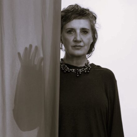 Imagem. Início da descrição de imagem: Retrato frontal em tom sépia de uma mulher de pele clara e cabelos curtos, cacheados e volumosos, predominantemente brancos. Ela olha diretamente para a câmera com uma expressão serena e um leve sorriso. Veste uma blusa escura de gola redonda e mangas compridas, e usa um colar largo e texturizado no pescoço. A artista está posicionada à direita, parcialmente oculta por um tecido translúcido claro que ocupa o lado esquerdo da composição. Através do tecido, vê-se a silhueta desfocada de uma mão aberta, criando um jogo de transparência e sobreposição de formas que mimetiza o estilo de suas próprias criações cênicas e visuais. O fundo à direita é liso e claro. Fim da descrição de imagem.