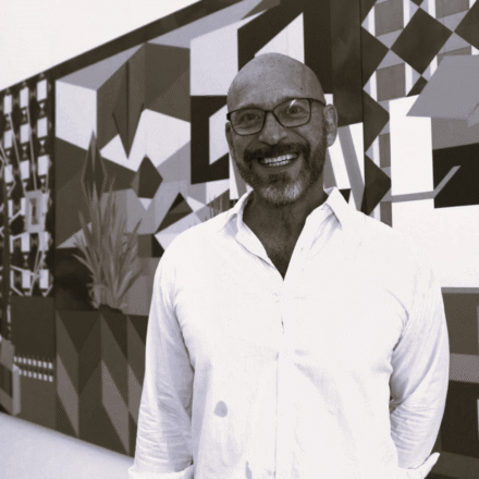 Imagem. Início da descrição de imagem: Retrato frontal em tom sépia de um homem careca, de pele clara, com barba e bigode grisalhos aparados. Ele sorri amplamente para a câmera, transmitindo uma expressão carismática. Usa óculos de grau com armação escura e veste uma camisa de botões clara, de tecido leve, com as mangas dobradas. O artista está posicionado em primeiro plano, à frente de um painel artístico de grandes dimensões que ocupa todo o fundo. A obra ao fundo é composta por complexas formas geométricas abstratas — triângulos, retângulos e polígonos — que criam uma ilusão de tridimensionalidade e profundidade arquitetônica, sugerindo um ambiente de galeria ou ateliê. Fim da descrição de imagem.