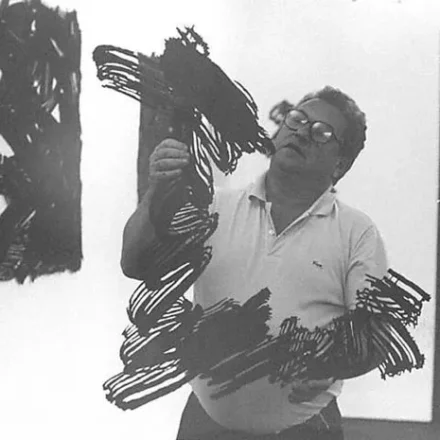 Imagem. Início da descrição de imagem: Fotografia em preto e branco de um homem de meia-idade, de pele clara e porte robusto. Ele olha para o alto enquanto manipula uma escultura de metal escura e vazada, composta por linhas sinuosas e hachuradas que mimetizam pinceladas gestuais no espaço. O artista usa óculos de grau com armação escura e veste uma camiseta polo clara. A cena ocorre em um ambiente interno iluminado: ao fundo, à esquerda, vê-se parte de outra obra de arte escura na parede; à direita, uma porta de vidro com moldura escura. A imagem captura o artista em um momento de interação direta com sua criação, destacando a materialidade e a escala de seu trabalho. Fim da descrição de imagem.