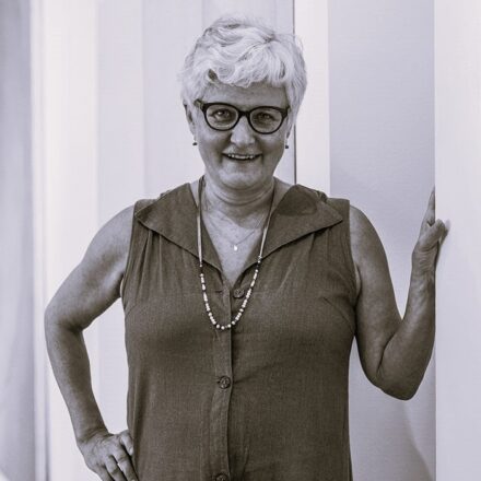 Imagem. Início da descrição de imagem: Retrato frontal em plano médio e tom sépia de uma mulher de meia-idade com pele clara, cabelos curtos, cacheados e brancos. Ela olha diretamente para a câmera com um sorriso aberto e gentil. Usa óculos de grau com armação escura e arredondada. Veste um macacão ou vestido sem mangas, com botões frontais e gola em "V", e usa dois colares: um curto com um pequeno pingente e outro mais longo com contas claras e escuras. A artista está posicionada entre o que parecem ser colunas ou divisórias claras de uma galeria; sua mão direita está apoiada na cintura e a mão esquerda encostada na coluna à direita, criando uma pose descontraída e acolhedora. Fim da descrição de imagem.