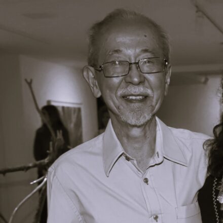 Imagem. Início da descrição de imagem: Retrato frontal em plano médio e tom sépia de um homem idoso de ascendência oriental. Ele tem cabelos claros raleados, barba e cavanhaque grisalhos, e olha diretamente para a câmera com um sorriso aberto e gentil. Usa óculos de grau com armação escura e retangular. Veste uma camisa social clara de mangas, com um bolso no peito. O artista está posicionado em um ambiente de galeria; ao fundo, de forma desfocada, observa-se uma figura feminina e o que parece ser uma instalação artística com elementos orgânicos e lineares, sugerindo o contexto de uma exposição. A iluminação é frontal, destacando a expressão serena e acolhedora do retratado. Fim da descrição de imagem.