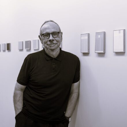 Imagem. Início da descrição de imagem: Retrato frontal em preto e branco (ou tom sépia) de um homem idoso de pele clara, calvo e com barba e bigode grisalhos. Ele está em pé, olha diretamente para a câmera com um sorriso aberto e espontâneo. Usa óculos de grau com armação escura e arredondada. Veste uma camisa polo escura com gola e botões, e calças escuras, com as mãos nos bolsos, e usa um relógio de pulso claro no braço esquerdo. O artista está posicionado centralizado à frente de uma parede branca onde estão penduradas uma série de pinturas abstratas emolduradas e dispostas em uma linha horizontal, que remetem a planos de cor e luz, típicas de suas pesquisas pictóricas. O chão é de cimento liso, sugerindo um ambiente de galeria ou museu. Fim da descrição de imagem.