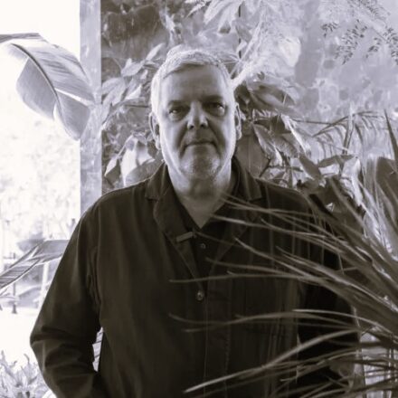 Imagem. Início da descrição de imagem: Retrato frontal em tom sépia do artista Caio Reisewitz. Ele é um homem de pele clara, cabelos curtos e grisalhos, e olha diretamente para a câmera com uma expressão serena. Veste uma camisa de botões escura. O artista está posicionado em meio a uma vegetação densa, com grandes folhas de plantas tropicais em primeiro plano e ao fundo, criando uma composição que remete à temática ambiental presente em sua obra fotográfica. Fim da descrição de imagem.