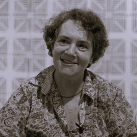 Imagem. Início da descrição de imagem: Retrato frontal em plano médio e tom sépia de uma mulher de meia-idade, de pele clara, com cabelos escuros, curtos e cacheados. Ela olha para o lado esquerdo da câmera com um sorriso aberto e espontâneo. Veste uma camisa estampada com folhas estilizadas e detalhadas e usa um colar fino e escuro no pescoço. A artista está posicionada à frente de uma parede com um padrão geométrico repetitivo de quadrados claros, que criam um efeito visual de azulejaria ou papel de parede, remetendo diretamente à sua pesquisa estética sobre ornamentação e padronagem. Fim da descrição de imagem.