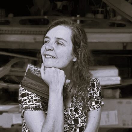 Imagem. Início da descrição de imagem: Retrato frontal em plano médio e tom sépia de uma mulher de pele clara, com longos cabelos ondulados e claros que caem sobre o ombro esquerdo. Ela olha para o lado esquerdo da câmera com uma expressão serena e um leve sorriso. Sua mão direita está posicionada abaixo do queixo, com o dedo indicador e o polegar apoiando o rosto em um gesto clássico de pensadora. Veste uma blusa estampada com uma gola geométrica detalhada. A artista está sentada em um ambiente interno onde o fundo, desfocado, sugere um ateliê ou espaço de criação, com formas que remetem a molduras, telas e talvez ferramentas de trabalho. Fim da descrição de imagem.