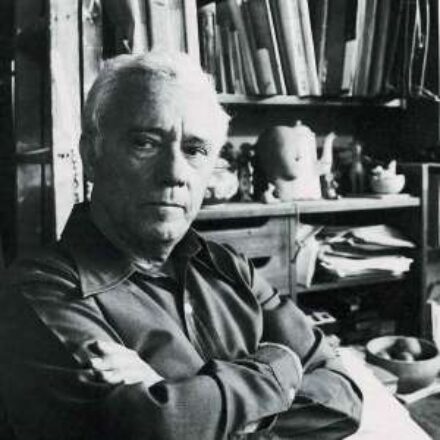 Imagem. Início da descrição de imagem: Retrato em preto e branco de um homem idoso de pele clara, com cabelos brancos e curtos. Ele olha diretamente para a câmera com uma expressão séria e braços cruzados na altura do peito. Veste uma camisa de mangas compridas em tom escuro. O artista está sentado à frente de uma estante de madeira repleta de livros, papéis e pequenos objetos cerâmicos, sugerindo seu ambiente de ateliê ou escritório pessoal. A composição é densa e revela um espaço de trabalho íntimo e intelectual. Fim da descrição de imagem.