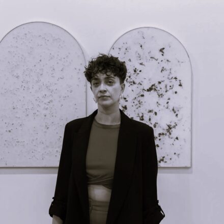 Imagem. Início da descrição de imagem: Retrato frontal em tom sépia de uma mulher de pele clara, com cabelos escuros, curtos e cacheados. Ela olha diretamente para a câmera com uma expressão serena e decidida. Veste um blazer escuro aberto sobre um top curto (cropped), revelando parte do abdômen, e calças de cintura alta. Usa brincos de argola claros. A artista está posicionada centralizada à frente de duas obras de arte em formato de arco (ogivais), fixadas em uma parede branca. As obras possuem uma textura interna salpicada de elementos escuros sobre um fundo claro, sugerindo o uso de materiais orgânicos ou minerais. Fim da descrição de imagem.