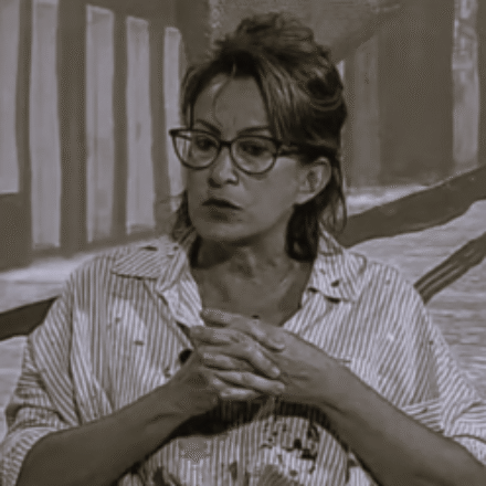 Imagem. Início da descrição de imagem: Retrato em tom sépia de uma mulher de pele clara, com cabelos escuros e lisos em um coque alto e despojado. Ela usa óculos de grau com armação escura em estilo "gatinho". Veste uma camisa de botões com listras verticais finas, que apresenta algumas manchas escuras (possivelmente tinta), sugerindo um momento de trabalho em ateliê. A artista está com as mãos entrelaçadas à frente do corpo e olha para o lado esquerdo com uma expressão atenta e pensativa. O fundo é composto por uma pintura de tons acinzentados com formas verticais que lembram janelas ou colunas, mantendo uma atmosfera de sobriedade e foco na figura. Fim da descrição de imagem.