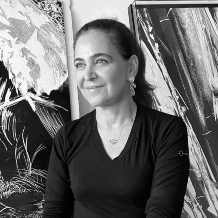 Imagem. Início da descrição de imagem: Retrato frontal em plano médio e em preto e branco de uma mulher de pele clara, com cabelos lisos e escuros presos para trás em um coque ou rabo de cavalo. Ela olha para o lado esquerdo da câmera com uma expressão serena e um leve sorriso. Usa brincos pendentes de metal e uma corrente fina com um pequeno pingente no pescoço. Veste uma blusa escura de gola em "V" com mangas compridas. A artista está posicionada centralizada à frente de duas grandes pinturas abstratas que ocupam todo o fundo. As obras ao fundo, embora em preto e branco na foto, exibem texturas espessas, sobreposições de camadas e pinceladas gestuais que conferem grande dinamismo e profundidade à composição, características da pesquisa matérica de Mignone. Fim da descrição de imagem.