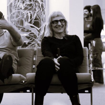 Imagem. Início da descrição de imagem: Retrato frontal em tom sépia de uma mulher de pele clara, com cabelos loiros curtos e franja reta. Ela usa óculos de grau com armação escura e grossa e olha para a esquerda da câmera com um sorriso largo e expressivo. A artista está sentada em uma poltrona de design moderno, vestindo uma blusa e calças pretas. Ao fundo, à esquerda, vê-se parte de uma obra de arte abstrata com linhas claras sobre fundo escuro, e à direita, duas pessoas aparecem de forma desfocada, sugerindo o ambiente de uma galeria ou museu. Fim da descrição de imagem.