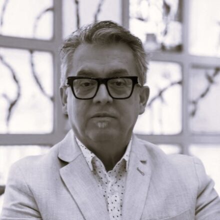 Imagem. Início da descrição de imagem: Retrato frontal em tom sépia de um homem de pele clara, com cabelos curtos e grisalhos penteados para o lado. Ele olha diretamente para a câmera com uma expressão séria. Usa óculos de grau com armação grossa e escura e possui um pequeno cavanhaque logo abaixo do lábio inferior. Veste um paletó de cor clara sobre uma camisa branca com pequenas estampas. Ao fundo, de forma desfocada, vê-se uma estrutura de vitrais com desenhos quadriculados com silhuetas de galhos de árvores. Fim da descrição de imagem.