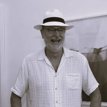 Imagem. Início da descrição de imagem: Retrato frontal em plano médio e em preto e branco de um homem idoso de pele clara, com barba e bigode grisalhos e densos. Ele olha para o lado direito da câmera com um sorriso aberto, espontâneo e radiante. Veste um chapéu panamá claro com uma fita escura larga na base, óculos de grau arredondados e uma camisa social clara estampada com um padrão xadrez miúdo de linhas escuras sobre fundo claro, de mangas curtas. No pescoço, carrega um cordão com pendentes. O artista está posicionado em primeiro plano, à frente de um fundo liso e claro; à direita, observa-se uma obra de arte abstrata em tons de cinza com formas fluidas e texturizadas, emoldurada em branco. Fim da descrição de imagem.