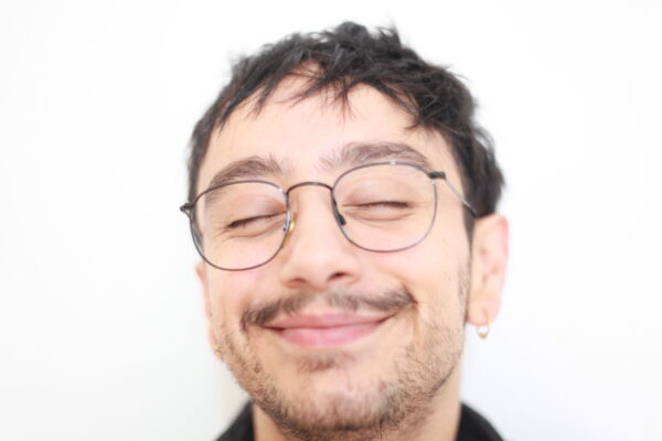 Imagem. Descrição da imagem: Um close-up do rosto de um homem jovem com olhos fechados e um sorriso satisfeito. Ele tem cabelos escuros e bagunçados e uma barba rala e não aparada. Ele está usando óculos de metal redondos e pequenos brincos de argola nas orelhas. O fundo é branco e está fora de foco. Fim da descrição.
