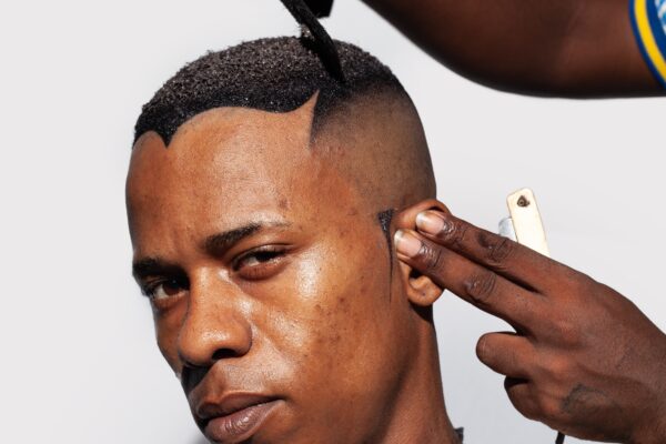 Imagem. Descrição de imagem: Um close médio de um homem negro recebendo um corte de cabelo contra um fundo branco e liso. Ele está olhando para a frente com uma expressão séria. Seu cabelo é preto e texturizado, e está sendo modelado com uma navalha e um pente. Em primeiro plano, o homem usa uma capa de barbeiro estampada com notas de dólar reais e desbotadas. O lado direito do rosto e a lateral da cabeça estão barbeados, revelando sua pele. Ele tem textura de pele natural, com poros e algumas marcas. Seus olhos são castanhos. Duas mãos de pele negra são visíveis no quadro. A mão esquerda segura uma navalha de cabo bege e branco perto de seu ouvido, finalizando o design do cabelo, enquanto a mão direita segura um pequeno pente preto e um objeto pequeno e preto na parte superior da cabeça, finalizando o visual. A mão direita também usa uma pulseira rosa brilhante e texturizada, e um detalhe de uma estampa azul e branca é visível em sua manga. A iluminação é clara e direta, vindo da frente, projetando uma sombra suave ao redor do pescoço e ombros. A foto é focada no rosto e no cabelo do homem, com as mãos e a capa borradas em segundo plano. Fim da descrição.