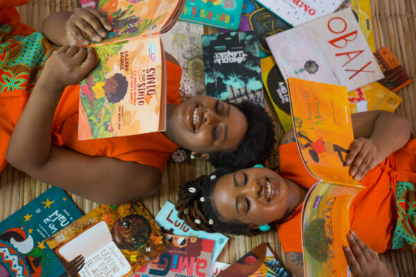 Imagem. Descrição da imagem: Duas mulheres negras estão deitadas de costas sobre uma esteira de palha clara, com as cabeças próximas ao centro e os corpos em direções opostas. Elas sorriem alegremente enquanto seguram livros abertos acima de seus rostos. Ambas possuem pele retinta e usam roupas de um laranja vibrante com detalhes em estampas africanas coloridas em tons de azul, amarelo e verde. A mulher à esquerda tem cabelos curtos e crespos, estilo black power, e usa brincos de argola brancos. O livro que ela segura tem o título 