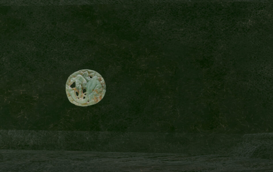 Imagem. Descrição da imagem: Close-up de uma parede de textura orgânica verde-escura em um ambiente virtual. Fixado à parede, há um disco circular esculpido em um material que lembra jade ou pedra esverdeada. O disco possui entalhes detalhados que formam figuras sinuosas e orgânicas. Fim da descrição da imagem.