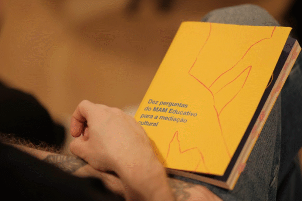 Imagem. Descrição da imagem: A foto apresenta um plano detalhado e em ângulo diagonal de uma pessoa segurando um pequeno livro amarelo sobre o colo. A pessoa possui pele clara e o antebraço esquerdo, que repousa sob o livro, exibe uma tatuagem escura e detalhada. Ela veste uma blusa preta e calça jeans azul. O livro possui uma capa amarela vibrante com desenhos de linhas finas e irregulares na cor vermelha, que sugerem formas abstratas ou contornos de montanhas. No canto inferior esquerdo da capa, lê-se o título em letras azuis: 