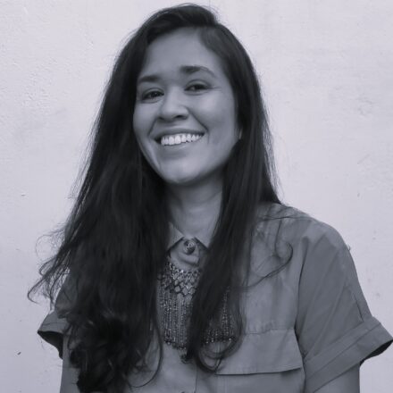 Imagem. Descrição da imagem: Um retrato fotográfico em preto e branco de Moara Tupinamba, uma mulher indígena com um sorriso radiante no rosto. Ela tem longos cabelos escuros e levemente ondulados que caem sobre os ombros, e sua pele tem uma textura natural e visível. Ela está de frente para a câmera, olhando diretamente para a lente com os olhos expressivos e brilhantes. Ela veste uma camisa de botão de mangas curtas e gola, feita de um tecido leve que parece algodão, e um colar de miçangas intrincado e texturizado pende no pescoço. O fundo é uma parede de estuque branca, ligeiramente texturizada, que cria um contraste limpo com a figura dela. A iluminação é suave e uniforme, sem sombras fortes, realçando a expressão alegre e a serenidade de sua presença. Fim da descrição.
