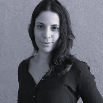 Imagem. Descrição da imagem: Fotografia em preto e branco de plano médio da curadora Juliana Caffé. Ela é uma mulher de pele clara, com cabelos escuros, levemente ondulados, longos e repartidos ao meio, que caem sobre seus ombros. Juliana encara a câmera com uma expressão serena e um leve sorriso fechado. Ela veste uma camisa escura de tecido leve, com botões pequenos aparentes na parte frontal. O fundo é uma parede de textura granulada e fosca, em um tom cinza médio, que cria um contraste suave com a figura. A iluminação é difusa, vindo da lateral esquerda, o que destaca suavemente os traços de seu rosto e a textura do seu cabelo. Fim da descrição.