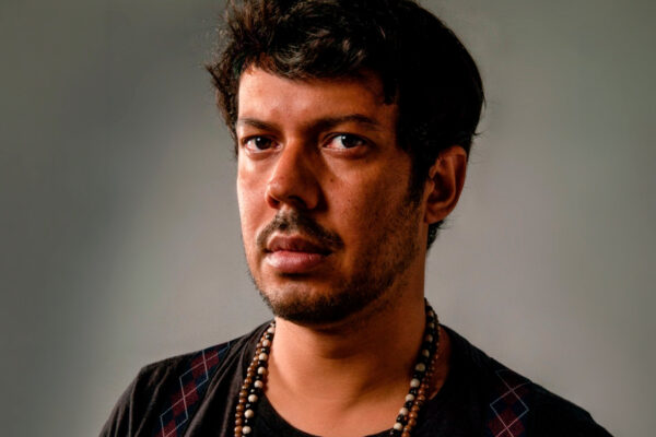 ## Descrição da Fotografia Imagem. Descrição da imagem: Um retrato de meio corpo de um homem jovem, de pele parda, posicionado do peito para cima contra um fundo cinza neutro e liso. Ele tem cabelos castanhos escuros, curtos e com uma textura levemente bagunçada no topo. Seu rosto apresenta uma barba rala e um bigode aparado; os olhos são escuros e ele mantém uma expressão séria e direta para a câmera. O homem veste uma camiseta preta básica e usa suspensórios com uma estampa de losangos (xadrez argyle) nas cores azul, vermelho e branco. No pescoço, carrega um colar longo composto por contas circulares em tons de marrom, bege e preto. A iluminação é dramática, vindo lateralmente, o que destaca a textura da sua pele e cria sombras suaves no lado esquerdo do rosto, conferindo profundidade ao registro. Fim da descrição.