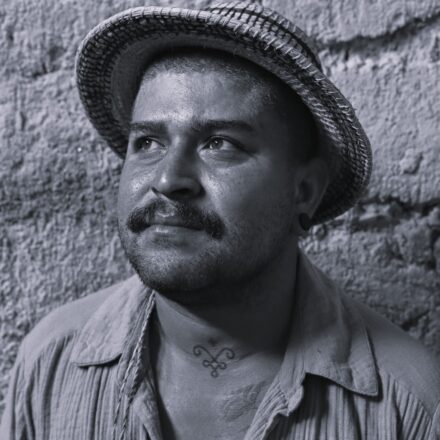 Imagem. Descrição da imagem: Uma fotografia em preto e branco, em primeiro plano e ângulo frontal, destaca o busto de um homem de pele parda posicionado do centro para a esquerda. Ele está com o rosto ligeiramente voltado para a esquerda e olha para o alto com uma expressão contemplativa. O homem tem cabelos muito curtos, sobrancelhas expressivas, olhos escuros e um bigode espesso acompanhado por uma barba rala e por fazer. Ele usa um chapéu de palha trançada com aba curta e levemente curvada para cima, um pequeno alargador ou brinco escuro no lóbulo da orelha esquerda visível e uma camisa de tecido rústico, amassado e listrado, com gola aberta. No centro do seu pescoço, há uma pequena tatuagem com um símbolo geométrico e simétrico de linhas curvas. O fundo da imagem é preenchido inteiramente por uma parede rústica de textura áspera e irregular, que se assemelha a reboco grosso, barro ou pedra. A iluminação é direcionada e vem do lado superior esquerdo, destacando o brilho nos olhos do homem, o relevo texturizado da parede ao fundo, os detalhes trançados do chapéu e as dobras de sua camisa. Fim da descrição.