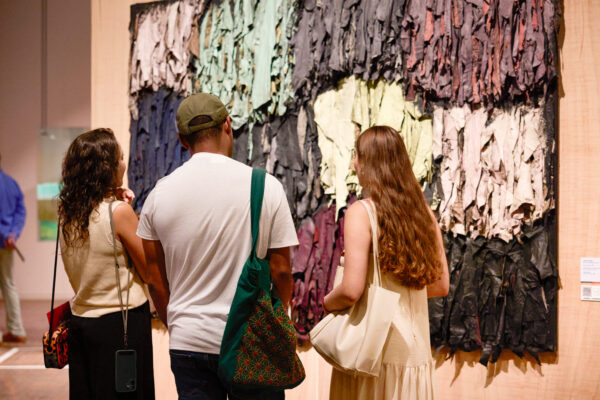Imagem. Descrição da imagem: Três pessoas estão de costas em primeiro plano, observando uma grande obra de arte abstrata e de textura marcante fixada em uma parede de tom claro. À esquerda, está uma mulher de pele clara e cabelos castanhos, cacheados e longos. Ela veste uma blusa regata de tricô na cor creme e calça preta. Traz a tiracolo uma pequena bolsa estampada em tons de laranja e preto e, pendurado por um cordão, um celular com capa escura. No centro, um homem de pele parda veste camiseta branca, calça jeans escura e um boné verde-oliva. Ele carrega uma grande bolsa de tecido verde com bordados coloridos no ombro esquerdo. À direita, está outra mulher de pele clara, com cabelos castanhos claros, ondulados e muito longos, que chegam até a cintura. Ela veste um vestido claro com alças finas e carrega uma bolsa de ombro bege. A obra de arte à frente deles é um painel retangular composto por várias tiras e pedaços de tecido ou couro franzidos e sobrepostos, criando uma textura volumosa e tridimensional. As cores da obra são dispostas em blocos verticais e horizontais, misturando tons de cinza, verde-claro, vinho, bege e preto. A iluminação do ambiente é focada e quente, destacando os detalhes e as sombras da textura da obra. Ao fundo, do lado esquerdo, vislumbra-se outra pessoa de calça clara e camisa azul, parcialmente visível. Fim da descrição.