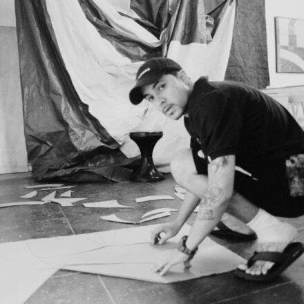 Imagem. Descrição da imagem: Uma fotografia em preto e branco, em plano médio e ângulo normal, mostra um jovem de pele clara agachado no chão de um estúdio ou ateliê de arte. Ele está posicionado do centro para a direita da imagem, de perfil para o lado esquerdo, mas com o rosto virado diretamente em direção à câmera. O jovem usa um boné escuro com aba virada para frente, camiseta escura de mangas curtas, bermuda escura, meias brancas e chinelos de dedo escuros. Ele tem cabelos curtos que aparecem sob o boné, olhos escuros, bigode e cavanhaque finos. No seu braço esquerdo, que está estendido em direção ao chão, há tatuagens visíveis, incluindo uma com o desenho de uma caveira. O jovem está agachado sobre os calcanhares e apoia as duas mãos sobre uma grande placa de papelão disposta no chão à sua frente. Ao redor e atrás dele, há vários pedaços recortados de papelão espalhados pelo piso escuro e liso. Ao fundo, no lado esquerdo, há uma grande lona ou tecido amassado e bicolor (com partes claras e escuras) pendurado na parede. No centro do plano de fundo, há um pequeno banco ou banqueta preta em formato de carretel. No lado direito, ao fundo, vê-se parte de uma bancada ou mesa comprida de madeira e, na parede branca mais distante, dois quadros retangulares emoldurados com artes abstratas pendurados lado a lado. A iluminação do ambiente é difusa e natural, vinda da lateral esquerda, criando sombras suaves no chão e nos objetos. Fim da descrição.