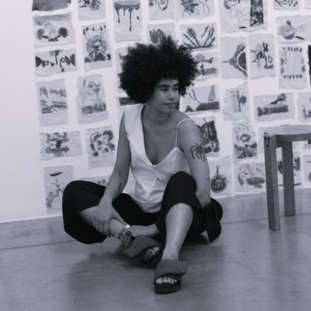 Imagem. Descrição da imagem: Uma fotografia em preto e branco, em plano médio e ângulo frontal, mostra uma jovem mulher negra de pele clara sentada no chão, no centro da composição. Ela tem um volumoso cabelo crespo em estilo crespo black power, sobrancelhas expressivas e olhos escuros voltados sutilmente para o lado direito da imagem com um leve sorriso. A mulher veste uma blusa clara e assimétrica: o lado direito possui uma gola ou alça larga dobrada e o lado esquerdo tem uma alça fina de estilo regata. Ela também usa calças pretas pantacourt largas e chinelos escuros de tiras grossas e acolchoadas. Em seu braço esquerdo, há uma tatuagem de uma palmeira ou coqueiro estilizado, e ela usa um colar delicado e anéis nos dedos. Ela está sentada de pernas cruzadas no chão cinza e liso de uma galeria ou ateliê. Ao fundo, uma grande parede branca está repleta de pequenos desenhos ou gravuras em preto e branco feitos em papel, dispostos em fileiras e colunas organizadas como um mosaico. No lado direito, em primeiro plano parcial, vê-se a perna e o assento de uma banqueta de madeira clara. A iluminação é suave e uniforme, destacando a textura do cabelo e os detalhes das obras de arte na parede. Fim da descrição.