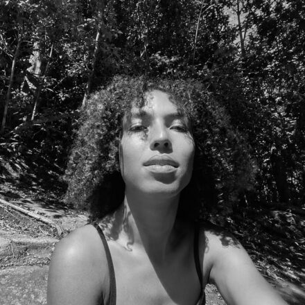 Imagem. Descrição da imagem: Uma fotografia em preto e branco, em primeiro plano e ângulo frontal levemente contra-plongée (de baixo para cima), foca no rosto e nos ombros de uma jovem mulher negra de pele clara. Ela está centralizada na imagem, com a cabeça levemente inclinada para trás e os lábios entreabertos, olhando em direção à câmera. Seu cabelo é crespo, volumoso, cheio de cachos definidos e está armado ao redor do rosto. Ela veste uma blusa escura de alças finas. O fundo da imagem exibe uma área de mata ou floresta densa com muitas árvores e folhagens que preenchem quase todo o espaço. Ela parece estar sentada em uma superfície rochosa ou inclinada no canto inferior esquerdo. A iluminação solar é intensa e direta, vinda de cima e do lado direito, projetando sombras nítidas das folhas e galhos das árvores sobre o lado esquerdo do seu rosto, pescoço e colo, criando um forte efeito de contraste de luz e sombra. Fim da descrição.