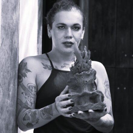 Imagem. Descrição da imagem: Uma fotografia em preto e branco, em plano médio e ângulo frontal, destaca uma mulher trans de pele clara, posicionada no centro da imagem, segurando uma escultura rústica com as duas mãos e olhando diretamente para a câmera. Ela tem cabelos escuros e crespos puxados e presos para trás, olhos delineados por maquiagem escura e batom marcante nos lábios. Veste uma blusa escura de alças finas estilo frente única. Seus braços estão repletos de tatuagens detalhadas, incluindo ilustrações de personagens e formas botânicas. No centro do colo, logo abaixo do pescoço, há uma tatuagem escrita em caixa alta com as palavras "CORPA EM TRÂNSITO" ou similar. Ela está ligeiramente encostada com o ombro direito em um batente ou coluna vertical de madeira clara no lado esquerdo da imagem. Ela segura uma escultura vertical de cerâmica ou argila texturizada, que possui pequenas perfurações e formas orgânicas que remetem a corais ou chamas. Ao fundo, no lado direito, há uma parede ou porta de madeira escura com tábuas verticais. A iluminação é direcionada, vinda do lado superior esquerdo, destacando o rosto, os ombros e os detalhes da peça esculpida em suas mãos. Fim da descrição.
