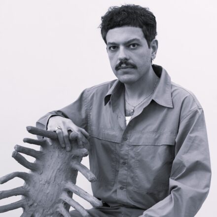 Imagem. Descrição da imagem: Um homem de pele clara, com cabelos pretos, curtos e ondulados, e um bigode marcante encara a câmera com uma expressão serena. Ele está posicionado do centro para a direita da imagem, vestindo uma camisa de botões de manga longa em um tom médio, levemente desabotoada no topo, revelando uma camiseta branca por baixo e um colar fino com um pequeno pingente. Ele também usa um brinco de argola pequena na orelha esquerda e um anel no dedo médio da mão direita. Sua mão direita repousa sobre uma escultura de argila texturizada que se projeta do canto inferior esquerdo. A peça possui um formato orgânico e alongado, assemelhando-se a uma espinha dorsal ou a uma criatura marinha, com várias ramificações pontiagudas que saem de um eixo central. O fundo é completamente liso e branco, destacando a figura do homem e a obra de arte. A foto é em preto e branco (escala de cinza), com uma iluminação suave e difusa que elimina sombras marcadas. Fim da descrição.