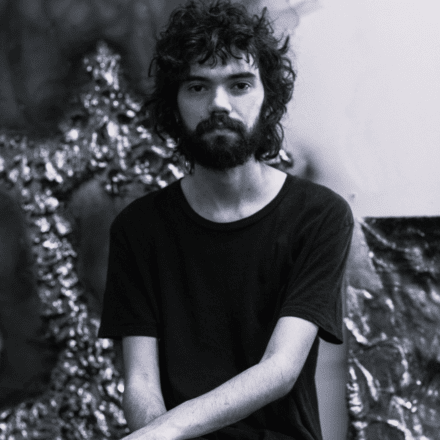 Imagem. Descrição da imagem: Um retrato em preto e branco, na vertical, mostra um jovem de pele clara centralizado na composição. Ele tem cabelos castanhos escuros, volumosos e bastante cacheados, que caem ligeiramente sobre a testa, e uma barba e bigode cheios e bem desenhados. Seus olhos são escuros e ele olha diretamente para a câmera com uma expressão serena e neutra. O homem veste uma camiseta preta básica de gola careca e mangas curtas. Seus braços estão cruzados à frente do corpo, apoiados sobre o colo, deixando à mostra seus antebraços magros. O fundo está ligeiramente desfocado, mas é possível notar elementos texturizados e brilhantes que lembram esculturas metálicas ou molduras ornamentadas e abstratas à esquerda e à direita, contrastando com uma parede de tom claro logo atrás dele. A iluminação é suave e frontal, destacando os detalhes do rosto e a textura dos cachos. Fim da descrição.