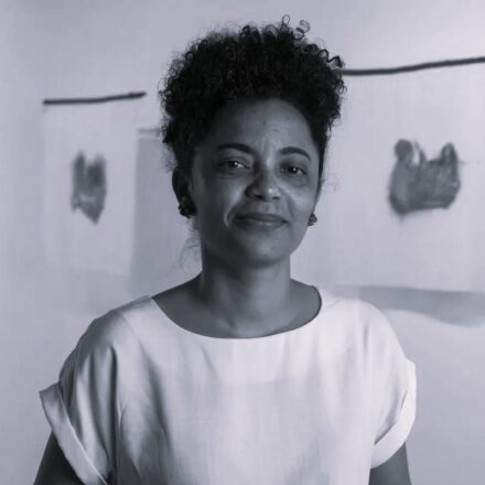 Imagem. Descrição da imagem: Uma mulher de pele parda e traços negros está centralizada em primeiro plano, olhando diretamente para a câmera com um sorriso discreto. Ela tem cabelos pretos, crespos e volumosos, no estilo "black power", que emolduram o topo de sua cabeça. Seus olhos são escuros e ela usa brincos pequenos. Veste uma blusa de tecido leve e cor clara com decote canoa e mangas curtas dobradas. A fotografia é em preto e branco. Ao fundo, o ambiente é uma galeria de arte ou exposição com paredes brancas e iluminação suave. Presas na parede atrás da mulher, há três obras de arte conceituais que parecem folhas de papel ou tecidos pendurados, contendo manchas escuras e abstratas no centro de cada uma. O fundo está levemente desfocado, mantendo o foco principal na mulher. Fim da descrição.