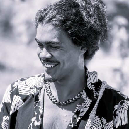 Imagem. Descrição da imagem: Um plano médio de um homem jovem de pele parda, sorrindo e olhando para baixo e para a sua direita. A foto está em preto e branco e a iluminação é natural e direta, vinda de cima, destacando suas feições. Ele tem cabelos escuros, volumosos, cacheados e presos em um coque alto. Possui sobrancelhas marcadas, olhos semicerrados pelo sorriso, bigode fininho e uma pequena linha de barba (cavanhaque) no queixo. Ele veste uma camisa de botão estampada com motivos florais e folhagens de tons contrastantes, que está aberta na frente, revelando parte do peito e o vislumbre de uma tatuagem na região do esterno. No pescoço, usa um colar feito de sementes esféricas ou contas artesanais em duas voltas. Uma alça de textura trançada ou de corda passa sobre o seu ombro esquerdo. O fundo da imagem está desfocado, sugerindo um ambiente externo gramado ou com vegetação sob a luz do dia. Fim da descrição.