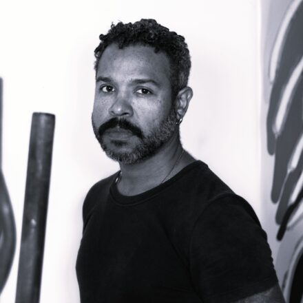 Imagem. Descrição da imagem: Retrato em preto e branco de um homem de pele retinta clara, posicionado do peito para cima, olhando diretamente para a câmera com uma expressão serena e focada. Ele tem cabelos curtos, crespos e cacheados, e uma barba cheia com bigode bem marcado, levemente grisalha nas laterais. O homem usa uma camiseta escura de gola careca, uma corrente fina no pescoço e um pequeno brinco de argola na orelha esquerda. A iluminação é suave e vem da esquerda, destacando as texturas de sua pele e da barba. O fundo é claro e minimalista; à esquerda, em primeiro plano e levemente desfocados, aparecem elementos verticais que parecem ser esculturas ou tubos, e à direita há parte de uma parede com grafismos ou pinturas abstratas em tons escuros. Fim da descrição.