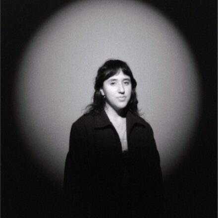 Imagem. Descrição da imagem: Retrato em preto e branco, com formato quadrado, focado no busto de uma mulher jovem de pele clara, posicionada no centro da composição. Ela tem cabelos escuros, levemente ondulados, na altura dos ombros e com uma franja reta sobre a testa. Seu semblante é sereno e ela direciona o olhar discretamente para o lado direito da câmera. Ela veste um casaco ou jaqueta escura e estruturada, de gola aberta. O fundo é composto por uma parede de textura fosca e granulada, iluminada por um forte feixe de luz circular, no estilo spotlight ou holofote, que destaca a jovem no centro e cria uma moldura de sombra escura e arredondada ao redor de toda a imagem. Fim da descrição.