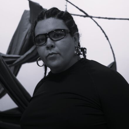 Imagem. Descrição da imagem: Um retrato em preto e branco foca no busto de uma pessoa que olha diretamente para a câmera com uma expressão séria e concentrada. Ela tem a pele com tom médio, cabelos escuros trançados rente à cabeça no estilo *cornrows* que terminam em pequenas tranças soltas nos ombros, e usa óculos de grau com armação escura e retangular. Na orelha visível à esquerda, há um brinco de argola metálica média e, na outra orelha, um brinco metálico mais grosso e arredondado. A pessoa veste uma blusa preta de manga longa e gola fechada. O fundo é claro e levemente acinzentado, composto por faixas de tecido escuro entrelaçadas que criam linhas diagonais e curvas abstratas ao redor da figura. Fim da descrição.
