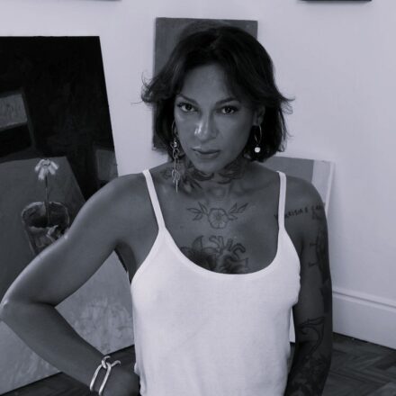 Imagem. Descrição da imagem: Uma fotografia em preto e branco, em plano médio, mostra uma mulher posicionada do centro para a esquerda da imagem, olhando diretamente para a câmera com uma expressão séria e marcante. Ela tem a pele com tom bronzeado médio, cabelos escuros, repicados e na altura do queixo, com textura levemente ondulada. A mulher usa uma regata branca de alças finas e decote arredondado. Seu corpo possui diversas tatuagens visíveis: um desenho orgânico detalhado cobrindo o pescoço, uma flor com folhas centralizada no colo e um coração anatômico logo abaixo. Seu braço esquerdo também apresenta várias tatuagens, incluindo escritas na altura do ombro. No pulso direito, ela usa um bracelete metálico espesso em formato de elos interligados e, nas orelhas, brincos grandes de argola pendentes, sendo o do lado esquerdo composto por argolas encadeadas. Ela está com a mão direita apoiada na cintura. O cenário ao fundo sugere um ateliê de arte ou estúdio com paredes claras e piso de taco de madeira. Há várias telas de pintura apoiadas no chão e encostadas nas paredes ao redor dela. À esquerda, uma grande tela escura mostra a pintura de um vaso com uma flor solitária. A iluminação é difusa e frontal, destacando os contornos de seu rosto, as tatuagens e o contraste entre as roupas e as obras de arte ao fundo. Fim da descrição.