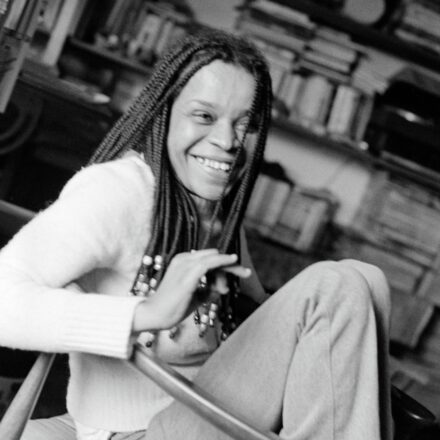 Imagem. Descrição da imagem: Uma fotografia em preto e branco, levemente inclinada, mostra a intelectual e ativista Lélia Gonzalez, uma mulher negra de pele clara, sorrindo de forma espontânea e calorosa. Ela está sentada em uma cadeira de madeira, com o corpo voltado ligeiramente para a esquerda, mas com o rosto virado em direção à câmera. Seu cabelo está penteado em tranças finas e longas (box braids) que caem sobre seus ombros, decoradas com pequenas miçangas de tons claros nas pontas. Lélia veste uma blusa de tricô de manga longa e cor clara, com punhos canelados, e calças compridas também claras. Ela está com as pernas dobradas na altura do peito, em uma postura descontraída, e sua mão direita está erguida na altura do peito, com os dedos levemente dobrados em um gesto dinâmico, como se estivesse conversando. O ambiente ao fundo está ligeiramente fora de foco, sugerindo um escritório ou biblioteca doméstica com prateleiras repletas de livros, papéis e objetos pessoais, criando uma atmosfera intelectual e acolhedora. A iluminação é suave e natural, destacando as texturas de sua pele, de suas roupas e os detalhes de seu sorriso marcante. Fim da descrição.