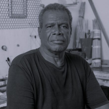 Imagem. Descrição da imagem: Um retrato em preto e branco, enquadrado do tórax para cima, mostra um homem negro de meia-idade posicionado no centro da imagem. Ele tem cabelos curtos, crespos e grisalhos, olhos escuros e uma expressão facial serena e compenetrada, olhando diretamente para a câmera. Ele veste uma camiseta preta básica de gola careca. O fundo está ligeiramente desfocado, sugerindo o ambiente de um ateliê ou oficina, onde se notam ferramentas, pincéis à esquerda, feixes de varas ou pincéis longos amarrados à direita, além de uma tela aramada na parede ao fundo. A iluminação é suave e vem de frente, destacando os detalhes de sua pele e a textura de suas roupas. Fim da descrição.