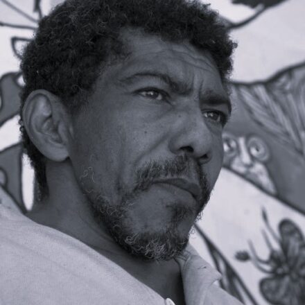 Imagem. Descrição da imagem: Um retrato em plano médio e em preto e branco de um homem de meia-idade, posicionado ligeiramente de perfil, olhando para o lado direito da imagem. Ele tem a pele retinta, cabelos curtos, crespos e escuros, e uma barba por fazer com fios grisalhos concentrados no queixo e no bigode. Sua expressão facial é séria e contemplativa, com marcas de expressão na testa e ao redor dos olhos. O homem veste uma camisa de gola polo de cor clara, com um botão visível perto do pescoço. O fundo da imagem está ligeiramente desfocado e mostra parte de um mural ou pintura com linhas artísticas, incluindo o que parece ser o desenho de uma borboleta e formas abstratas. A iluminação é natural e vem da direção para onde o homem olha, destacando os detalhes texturizados de sua pele e cabelo. Fim da descrição.
