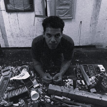 Imagem. Descrição da imagem: Uma fotografia em preto e branco com textura granulada mostra um homem de pele retinta clara e cabelos pretos crespos e curtos no centro da composição. Ele está posicionado atrás de uma bancada de trabalho, inclinado para a frente e olhando diretamente para a câmera com uma expressão séria. Seus braços estão apoiados na mesa e suas mãos manuseiam pequenos detritos. A bancada à sua frente está repleta de ferramentas de marcenaria e escultura, como uma plaina manual, um formão, uma escova de limpeza, um martelo e pedaços de madeira talhados, além de muitas raspas, lascas de madeira e um copo descartável espalhados pela superfície. Ao fundo, a parede texturizada e com marcas de desgaste exibe várias obras de arte abstratas e moldes pendurados. A iluminação vem de cima, destacando o topo da cabeça do homem e os objetos sobre a mesa, criando sombras marcadas no ambiente de atelier. Fim da descrição.
