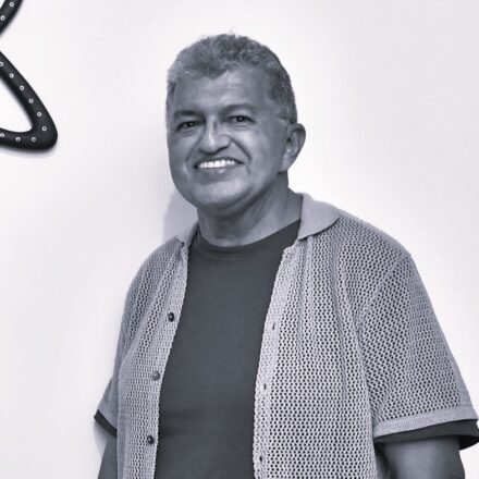 Imagem. Descrição da imagem: Um retrato em preto e branco, do plano médio para cima, mostra um homem de meia-idade sorrindo, posicionado ligeiramente à direita do centro. Ele tem a pele bronzeada, cabelos curtos, crespos e grisalhos, e olhos escuros. Seu sorriso é largo, exibindo os dentes superiores alinhados. Ele veste uma camiseta escura por baixo de uma camisa de mangas curtas aberta, feita de um tecido leve e texturizado com pequenos furos, semelhante a um tricô ou crochê. A camisa possui gola e botões pequenos na parte frontal esquerda. O fundo é uma parede clara e lisa, com uma iluminação suave que vem da frente e do alto, projetando uma leve sombra do homem na parede atrás dele. No canto superior esquerdo, aparece um detalhe parcial de um objeto escuro e sinuoso decorado com pequenos pontos claros alinhados, que lembram lâmpadas ou tachas. Fim da descrição.