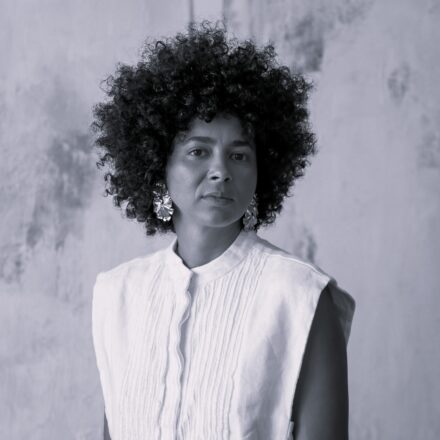 Imagem. Descrição da imagem: Retrato centralizado de uma mulher de pele parda e traços marcantes, fotografada da cintura para cima em um estilo monocromático (preto e branco). Ela tem um volumoso cabelo crespo, estilo afro, que emoldura perfeitamente o seu rosto. A sua expressão facial é séria, com o olhar direcionado de forma firme e profunda para a câmera. Ela veste uma blusa ou colete de cor clara (branca ou off-white) sem mangas, com gola padre curta e um detalhe plissado texturizado com linhas verticais na parte frontal. Em suas orelhas, destacam-se brincos grandes, metalizados e com formato orgânico e irregular, que reluzem discretamente. O fundo da imagem é uma parede com textura rústica e manchas abstratas em tons de cinza, criando um ambiente minimalista. A iluminação é suave e direcionada, destacando os contornos de seu rosto e a textura de seu cabelo. Fim da descrição.