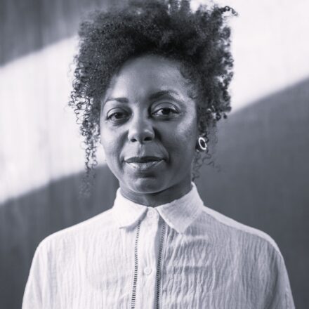 Imagem. Descrição da imagem: Retrato em plano médio, preto e branco, de uma mulher negra que olha diretamente para a câmera com uma expressão serena. Ela possui cabelos crespos, volumosos e texturizados, presos no estilo "afro puff" no topo da cabeça, com alguns cachos finos e soltos caindo suavemente pelas laterais do rosto. A textura de sua pele é lisa e exibe um brilho sutil nas áreas da testa, nariz e maçãs do rosto. Seus olhos são escuros e expressivos, e seus lábios estão suavemente fechados em um meio-sorriso discreto. Ela veste uma camisa social de cor clara, abotoada até o colarinho, com uma textura levemente amassada no tecido e um detalhe vertical de costura rendada que acompanha a linha dos botões centrais. Na orelha esquerda, é possível ver um pequeno brinco em formato de argola. O fundo da imagem é abstrato e está fora de foco, dividido diagonalmente por uma faixa clara e uma área mais escura, o que destaca a figura da mulher no centro. Fim da descrição.