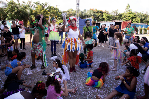 Imagem. Descrição da imagem: Em um plano amplo ao ar livre durante o dia, diversas pessoas e crianças vestindo roupas coloridas e fantasias divertem-se em uma praça pavimentada com pequenos paralelepípedos e confetes espalhados pelo chão. No centro da composição, destaca-se uma mulher de pele parda com maquiagem festiva, usando blusa regata branca e uma saia rodada cheia de penas coloridas em tons de azul, amarelo e vermelho. Ela ergue um instrumento musical com a mão direita. À sua esquerda, um homem de pele negra vestindo bermuda estampada e adornos verdes dança com os braços erguidos. Diversas crianças com saias de tutu, fantasias e laços no cabelo estão sentadas e em pé ao redor brincando no chão. Ao fundo, observam-se outras pessoas assistindo à apresentação e árvores sob a iluminação natural do sol. Fim da descrição.