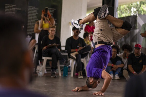 Imagem. Descrição da imagem: No centro da cena, um homem de pele clara com cabelos bem curtos executa uma manobra acrobática de breakdance, apoiando as mãos e a cabeça no chão enquanto mantém o corpo de ponta-cabeça e as pernas dobradas no ar. Ele veste camiseta roxa, bermuda cáqui, joelheiras pretas e tênis esportivos brancos com detalhes pretos e azuis. Ao fundo, em uma área ampla e iluminada por luz natural difusa, várias pessoas de diferentes tons de pele observam a performance sentadas em cadeiras e no chão. À esquerda, uma pessoa grava a cena com um celular; à direita, espectadores usam bonés e roupas casuais. Fim da descrição.