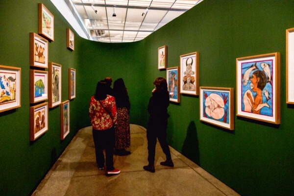 Imagem. Descrição da imagem: Em uma fotografia com ângulo amplo em perspectiva central, observa-se o corredor de uma galeria de arte com paredes altas pintadas de verde-escuro e piso de concreto polido. Nas paredes à esquerda e à direita, há diversos quadros coloridos e figurativos com molduras de madeira clara dispostos em diferentes alturas. No centro da composição, de costas para a câmera, três pessoas observam as obras expostas: à esquerda, uma pessoa de pele clara veste uma jaqueta vermelha estampada com flores, calça preta e tênis escuros com sola e cadarços brancos; ao centro, outra pessoa de pele clara e cabelos escuros compridos veste uma saia longa estampada; à direita, uma pessoa de pele clara e cabelos castanhos curtos veste calça e casaco pretos. A iluminação é focada e direta, projetada a partir de trilhos no teto alto e criando sombras marcadas das pessoas sobre a parede direita. Fim da descrição.